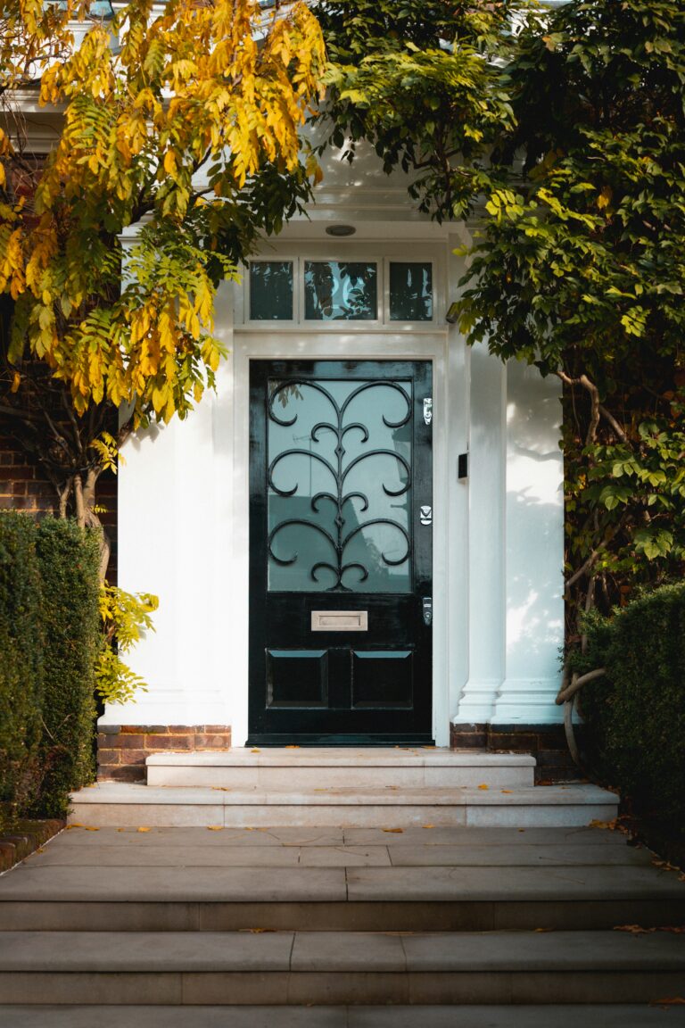 Elegant black door with decorative glass, framed by lush greenery and autumn leaves, welcomes visitors to a charming home entrance.
