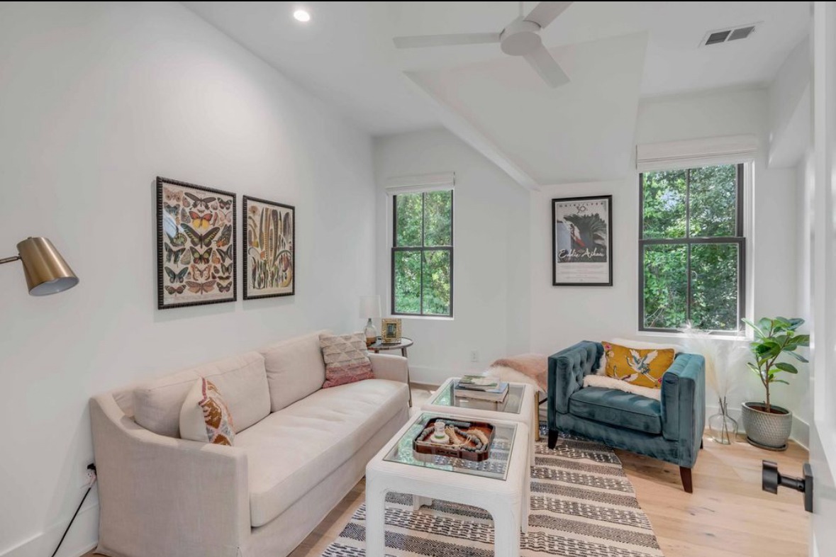 Bright, modern living room with a cream sofa, teal armchair, and nature-themed wall art. Large windows bring in natural light.
