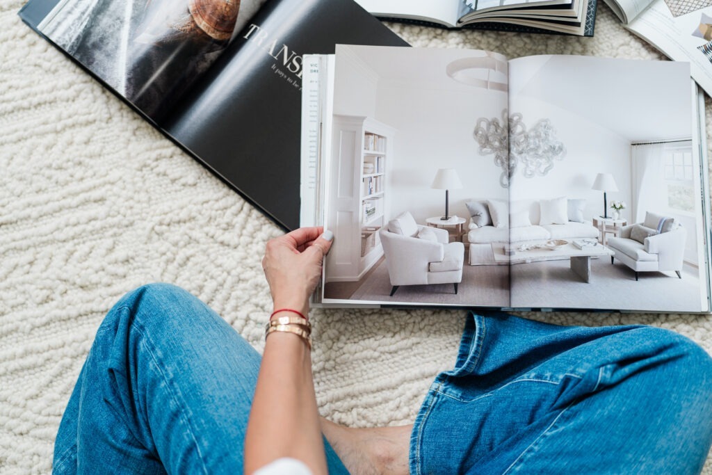 A person in a white shirt and blue jeans sits on a textured rug, holding an open design magazine featuring a modern, light-filled living room.