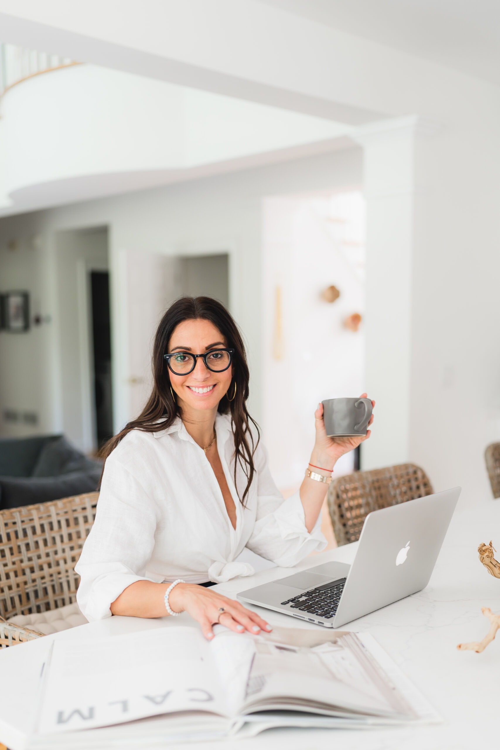 A woman in a white blouse sits at a table with a laptop and a magazine, holding a coffee cup and smiling, in a bright, modern workspace.