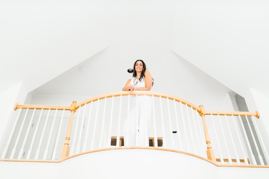 Woman in white attire stands on a wooden balcony, smiling down. Bright, airy space with high ceilings emphasizes a modern aesthetic.