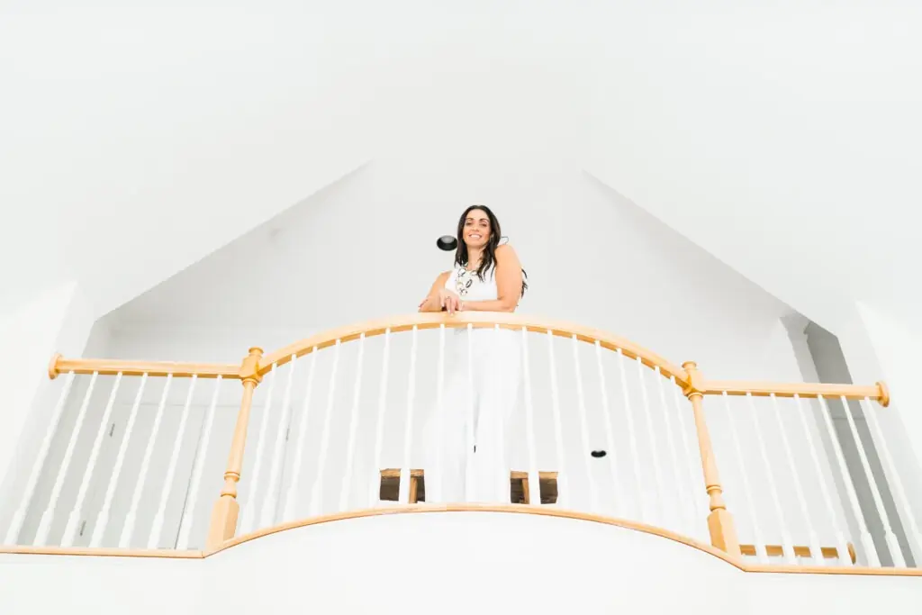 Woman in white attire stands on a wooden balcony, smiling down. Bright, airy space with high ceilings emphasizes a modern aesthetic.