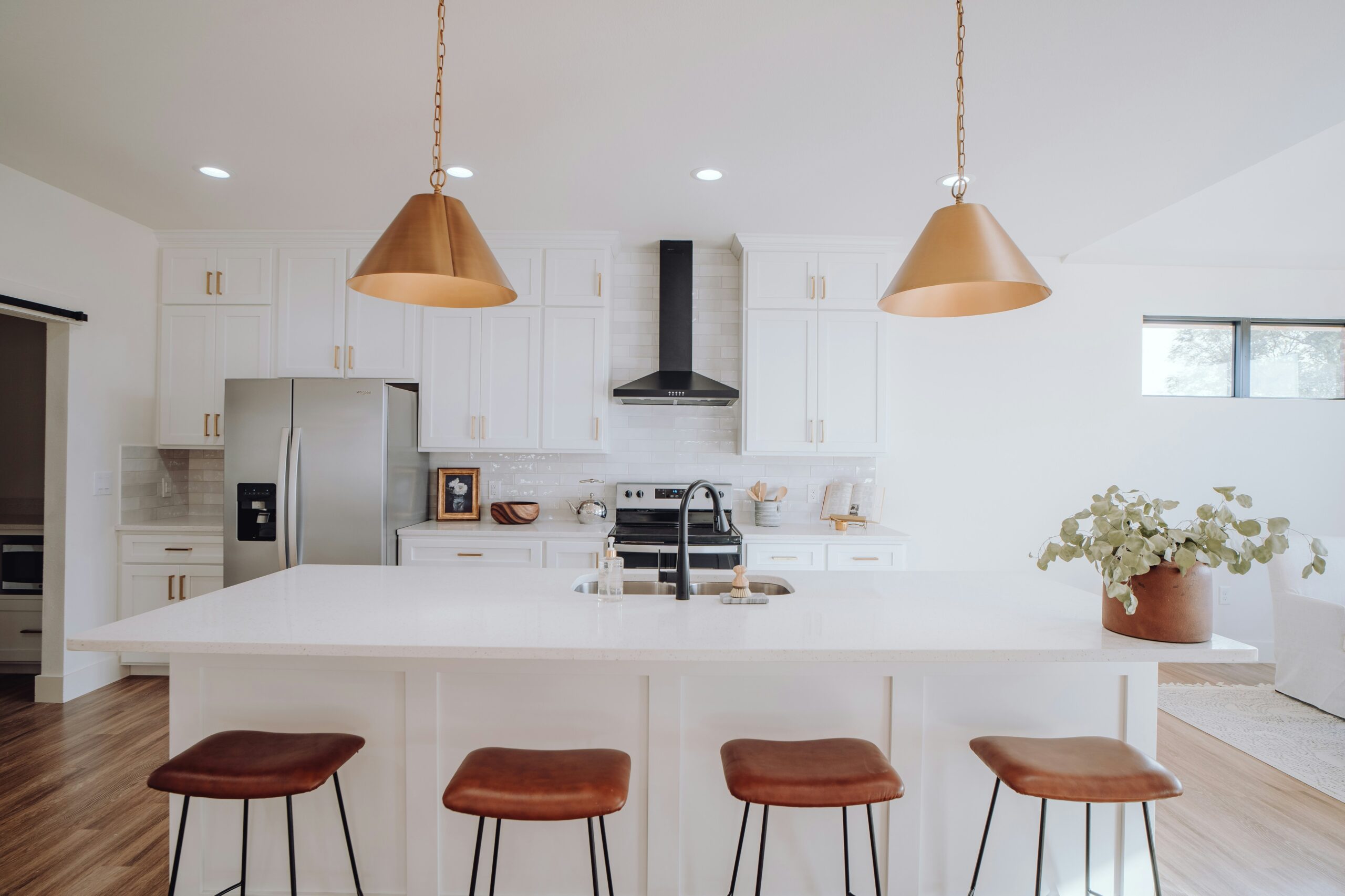 Modern kitchen featuring a large white island, brown leather bar stools, and stylish pendant lights. The clean design includes stainless steel appliances and a potted plant.