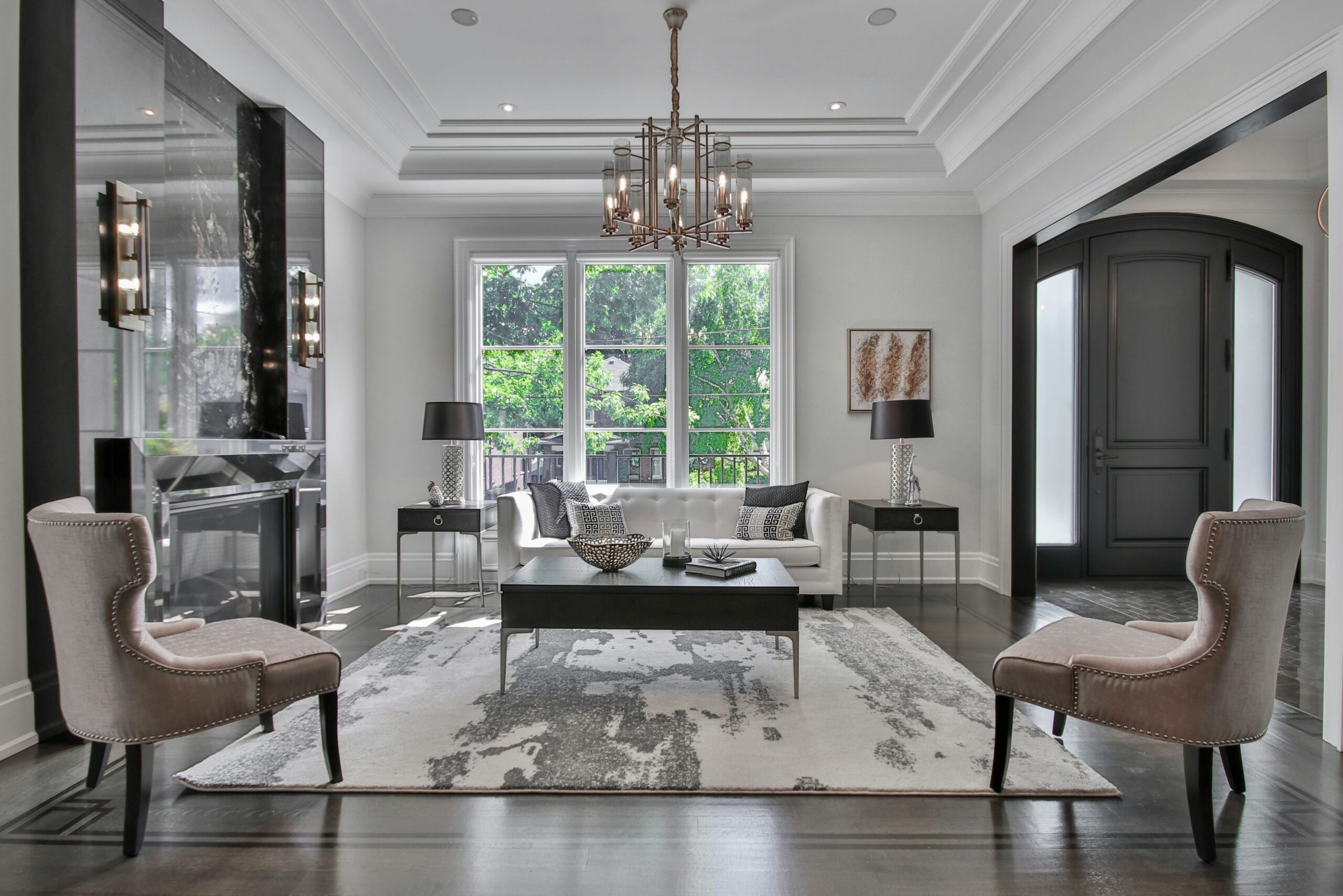 Elegant living room featuring a white sofa, black accent tables, and a stylish chandelier, illuminated by natural light from large windows.