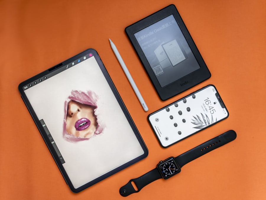 Digital art tools arranged on an orange background, featuring a tablet with artwork, a stylus, an e-reader, a smartphone, and a smartwatch, showcasing creative technology.